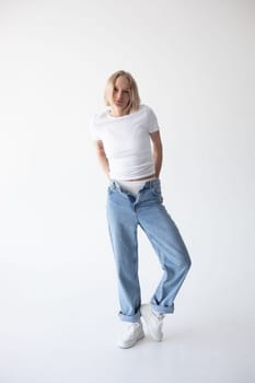 Beautiful blonde girl in a white T-shirt and blue jeans and sneakers posing on a white background. High quality photo