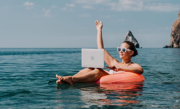 Woman freelancer works on laptop swimming in sea on pink inflatable ring. Pretty lady typing on computer while floating in the sea on inflatable donut at sunset. Freelance, remote work on vacation