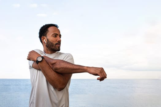 African American man stretching arm, warming up before workout and running session outdoors. Male exercising near the ocean. Health and fitness concept.