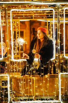 Woman holding sparkler night while celebrating Christmas outside. Dressed in a fur coat and a red headband. Blurred christmas decorations in the background. Selective focus.