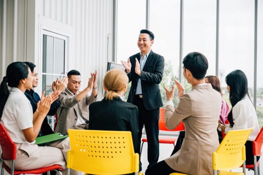 Asian man presents his work in meeting room receiving praise and compliments from colleagues. The audience claps with happiness celebrating his achievement. It signifies teamwork and business success.