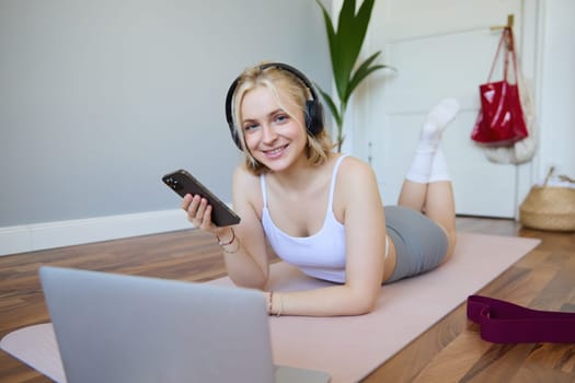 Portrait of young sporty woman, connects to online workout training session, doing exercises, lying on rubber mat, listens to fitness instructor in headphones, holding mobile phone.