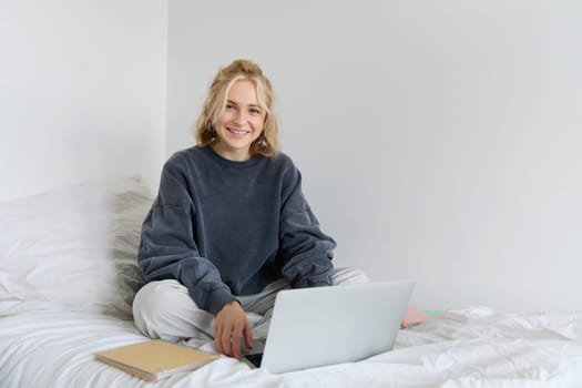 Portrait of young smiling woman studying in her bed, working from home in bedroom, sitting with laptop and notebooks on lotus pose, looking happy and relaxed.