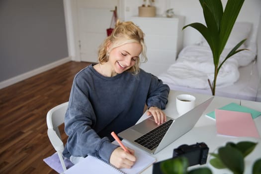 Portrait of smiling, beautiful blond woman, writing down notes, doing homework, studying from home, doing distance learning, online course, working remotely in her room.