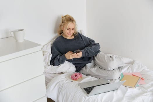 Portrait of smiling candid woman, lying in bed with doughnut, using smartphone and laptop, resting at home in bedroom, watching tv show or chatting online.