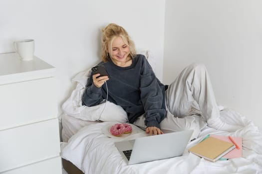 Portrait of smiling candid woman, lying in bed with doughnut, using smartphone and laptop, resting at home in bedroom, watching tv show or chatting online.