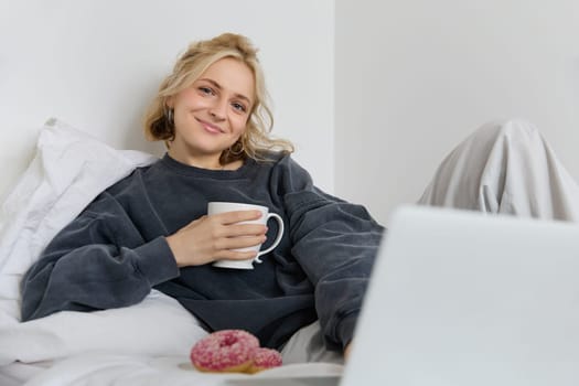 Portrait of happy woman enjoying day-off at home, lying in bed, watching tv show on laptop, looking at screen and smiling, drinking tea and eating in bedroom.