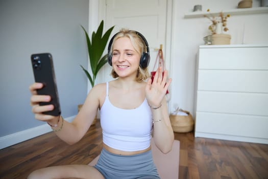Portrait of young sporty woman in headphones, records video on her smartphone, live streaming and saying hello to followers while doing workout training at home.