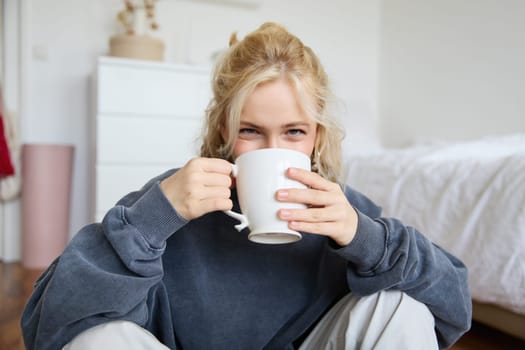 Portrait of young blond woman in casual clothes, sits on bedroom floor with cup of tea, drinking and looking at camera.