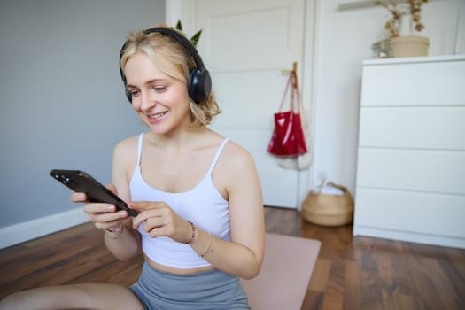 Portrait of young blond woman in headphones, turning on yoga, workout app on smartphone, choosing music on mobile phone application, sitting on rubber mat in room.