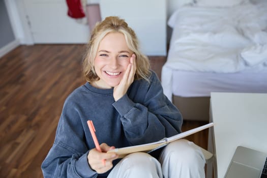 Portrait of smiling, charismatic young woman, writing down notes, making plans and putting it in planner, holding journal, sitting in bedroom and looking happy at camera.