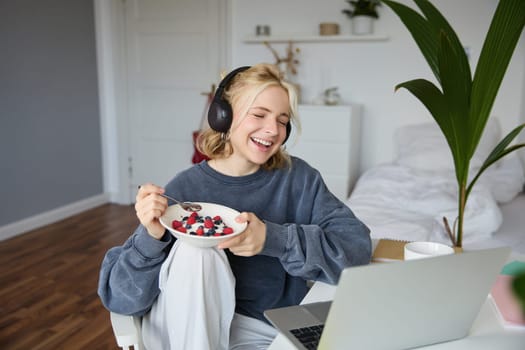 Portrait of smiling young woman, watching tv show in headphones, eating breakfast and looking at laptop screen.