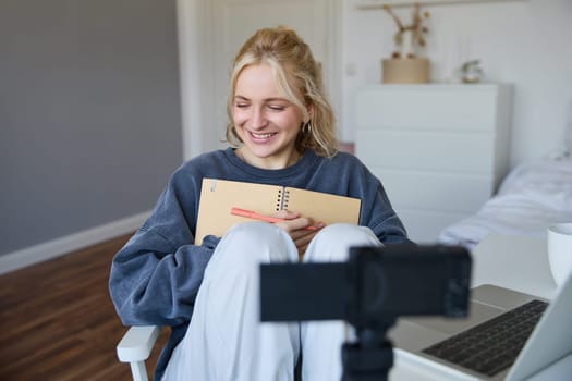 Portrait of smiling cute woman, lifestyle blogger, sits in her room with daily journal or planner, records video on digital camera, creates content for social media about daily routine.