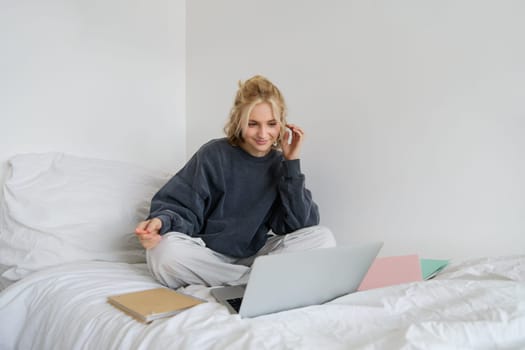 Portrait of female student, woman sits on bed with laptop and notebooks, studying online, remote education concept. Girl freelancer working from her bedroom.