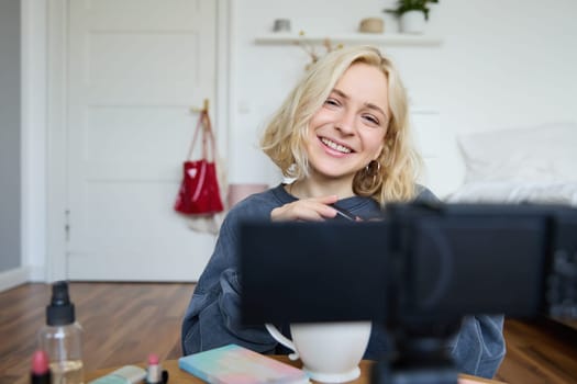 Close up portrait of young girl vlogger, teenager records video blog about makeup, uses camera on stabiliser, sitting on floor near coffee table, applying mascara and cosmetic products.