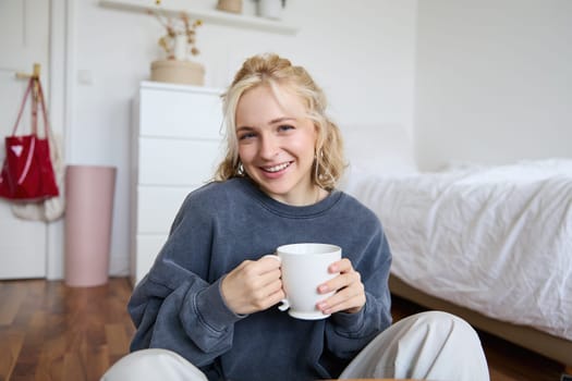 Portrait of young beautiful woman in casual clothes, sitting on bedroom floor with cup of tea, drinking and smiling, looking at camera, recording a lifestyle vlog.