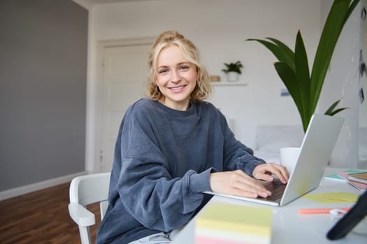 Portrait of smiling young woman, college student sits in her room, does homework, studies remotely from home, uses laptop for freelance work.