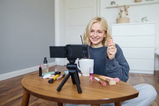 Portrait of beautiful, smiling blond woman, girl recording video of her makeup tutorial for social media, vlogger sitting on floor in her room, using stabiliser to create content, reviewing mascara.