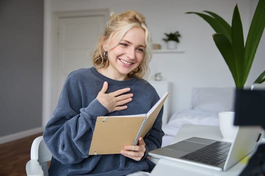 Portrait of young smiling blond woman, working from home, online chatting, using digital video camera, recording vlog, holding notebook, reading notes, explaining something.