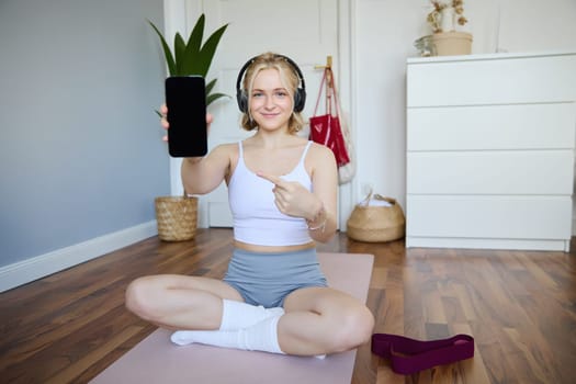 Portrait of smiling, fit and healthy, fitness instructor at home, pointing finger and showing her smartphone screen, recommending sports app for workout.