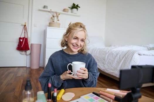 Portrait of beautiful social media beauty blogger, sitting in front of digital camera on floor in bedroom, drinking tea and chatting, talking to followers.