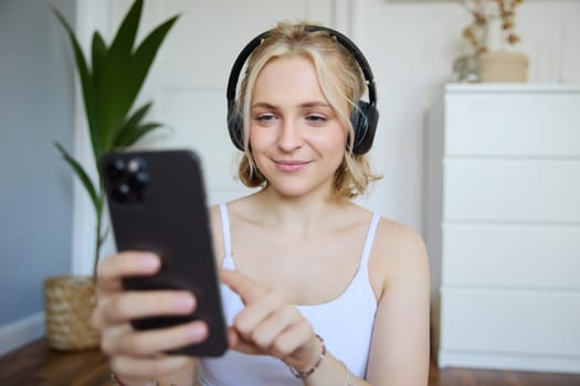 Close up portrait of smiling sporty woman, wearing headphones, using mobile phone, workout app while exercising at home. Wellbeing and lifestyle concept