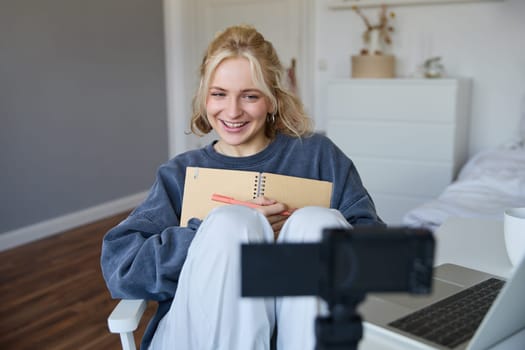 Portrait of cute, smiling young social media content creator, girl records video on digital camera and stabiliser, holds notebook, talks to audience, vlogging in her room.