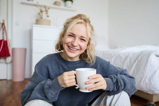 Portrait of young woman sitting on bedroom floor, drinking tea, holding white mug and smiling at camera.