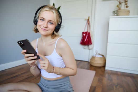 Portrait of smiling fitness woman, wearing wireless headphones, holding mobile phone, working out with sports app on smartphone, sitting on yoga rubber mat, exercising at home.