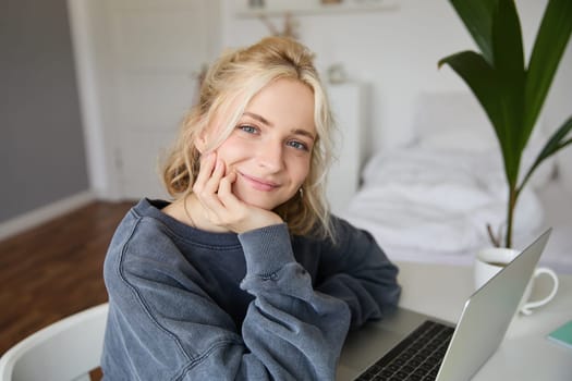 Portrait of young smiling woman, female student sitting in her room with laptop, looking cute at camera.