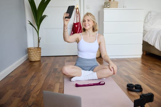 Portrait of stylish young woman, sitting on rubber mat on floor at home, taking selfie while doing workout, yoga session, posting photo on social media.