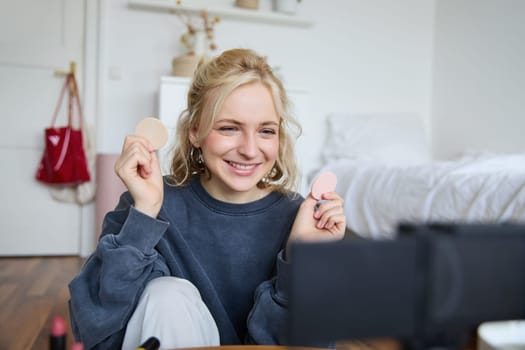 Portrait of young woman, beauty content creator, sitting in a room in front of digital camera, recording makeup tutorial vlog, showing cosmetic facial products.