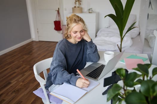 Lifestyle image of young woman writing down something in notebook, making notes, studying online, doing course in internet, listening to interesting information, using laptop.