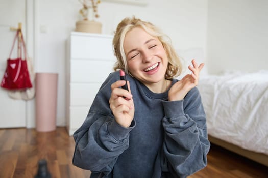 Portrait of smiling beautiful woman in her room, sitting and showing lipstick, recommending favourite beauty product, content maker recording a video of herself for social media blog.