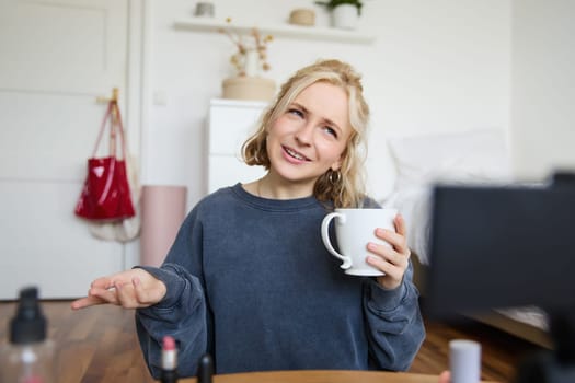 Portrait of blond young lifestyle blogger, woman records video of her talking about life and beauty, sits in front of camera, holds cup, drinks tea, does blog content for social media account.