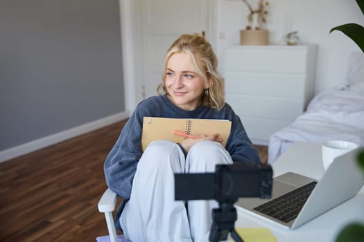 Social media and lifestyle content concept. Young happy smiling woman, sits in her room with notebook, talks at digital camera, having conversation with followers, does lifestyle vlog.