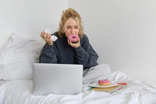 Portrait of sad, crying young woman, staying at home, sitting in bed with doughnut and comfort food, looking at something upsetting on laptop screen.