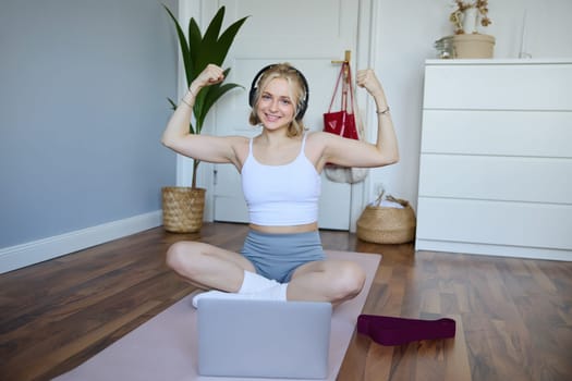 Portrait of young happy woman, fitness blogger in wireless headphones, working out at home, sits on rubber mat with laptop and wireless headphones, shows her muscles.