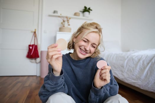 Portrait of happy, beautiful blond woman, showing makeup products on camera, recording vlog, social media content for followers, online live stream about beauty items.