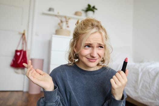 Portrait of woman, vlogger looking disappointed, showing lipstick and shrugging shoulders, recording video about makeup for social media account.