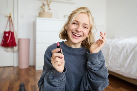 Portrait of young social media influencer, woman recording a video with beauty products, showing her makeup on camera, holding lipstick and smiling, sitting on floor.