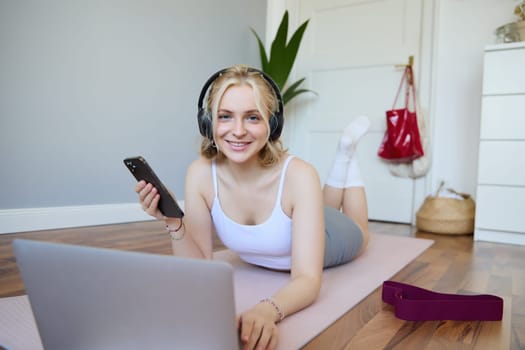 Portrait of happy blond woman in headphones, lying on rubber mat, listens to music in headphones after workout training session, using laptop.