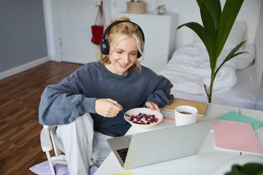 Portrait of happy young blond woman, sitting in a room, watching movie on laptop and eating healthy breakfast, drinking tea, resting on weekend.