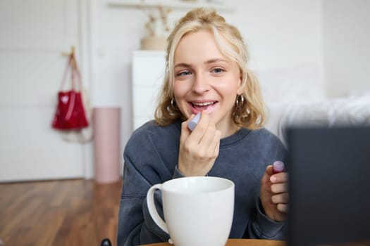 Lifestyle, beauty blogger, woman recording video of her putting on makeup, talking to camera, making online tutorial, showing her lip gloss or lipstick to followers, sitting on floor with cup of tea.