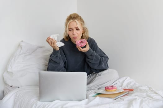 Portrait of woman watching sad show on laptop, eating doughnut and wiping tears with napkin, sitting on bed, spending time at home while being upset.