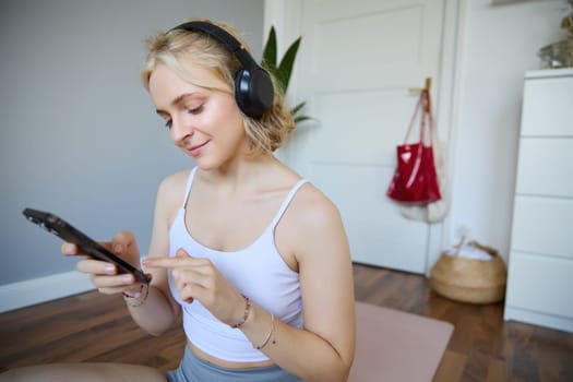 Portrait of young sporty woman in wireless headphones, sitting with smartphone on rubber mat, using workout training app, fitness application on her phone, listening in earphones.