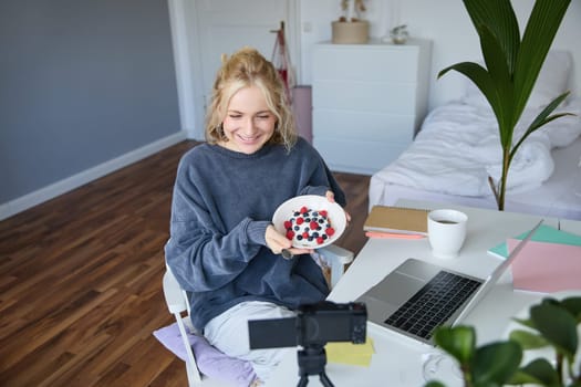 Portrait of cute young girl vlogger, showing her breakfast on camera, recording vlog in her bedroom, holding dessert, eating.