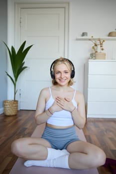 Portrait of woman feeling relaxed and in peace after meditation or yoga training at home, holding hands on chest, wearing headphones, smiling at camera.