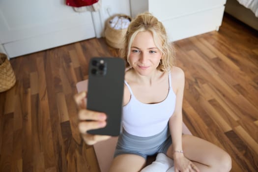 Young sporty fitness trainer, yoga instructor does live stream, takes selfies on smartphone while sitting on rubber mat at home, recording her workout training session.