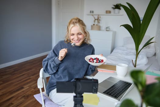 Portrait of smiling, candid young woman, content creator, eating bowl of dessert and looking at digital camera, recording vlog for followers on social media.